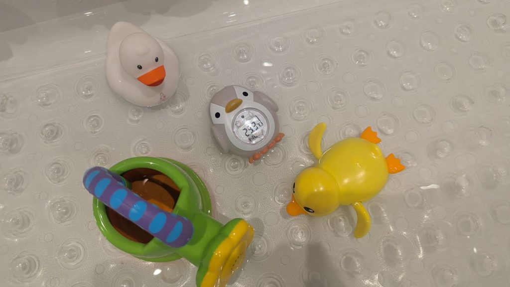 Bath toys including a rubber duck, watering can, and Nuby penguin digital thermometer floating on a white bath mat during a 10 month old bedtime routine.