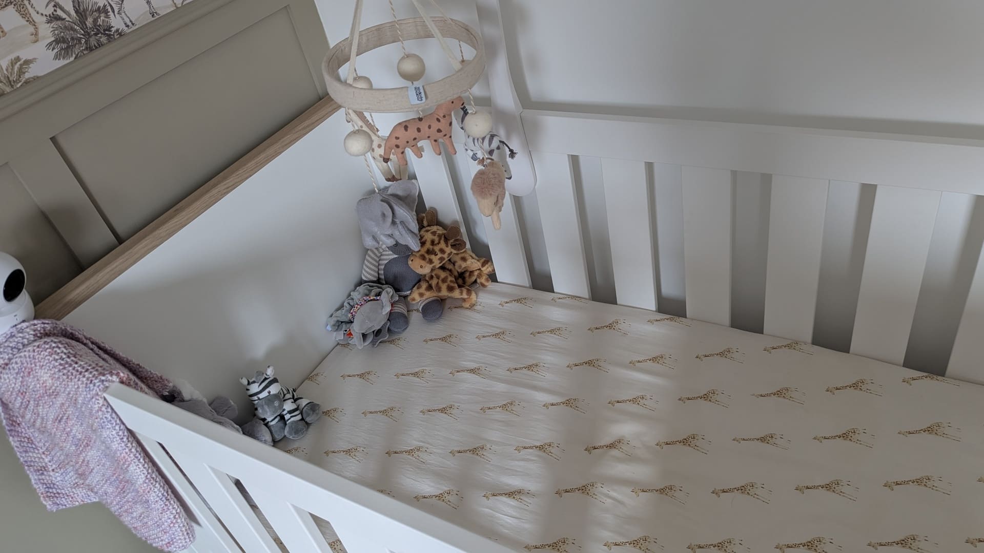 A white wooden cot with giraffe patterned bedding, plush toys, hanging mobile, and a baby monitor for a 10 month old bedtime routine.