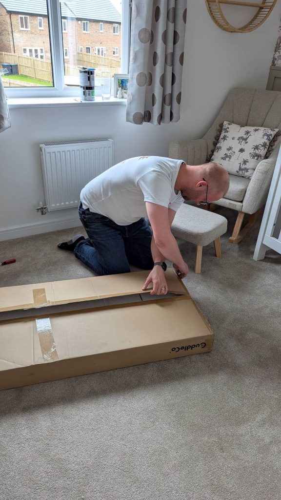 Dad kneeling on the carpet unboxing a large flat-pack cardboard box for nursery furniture.