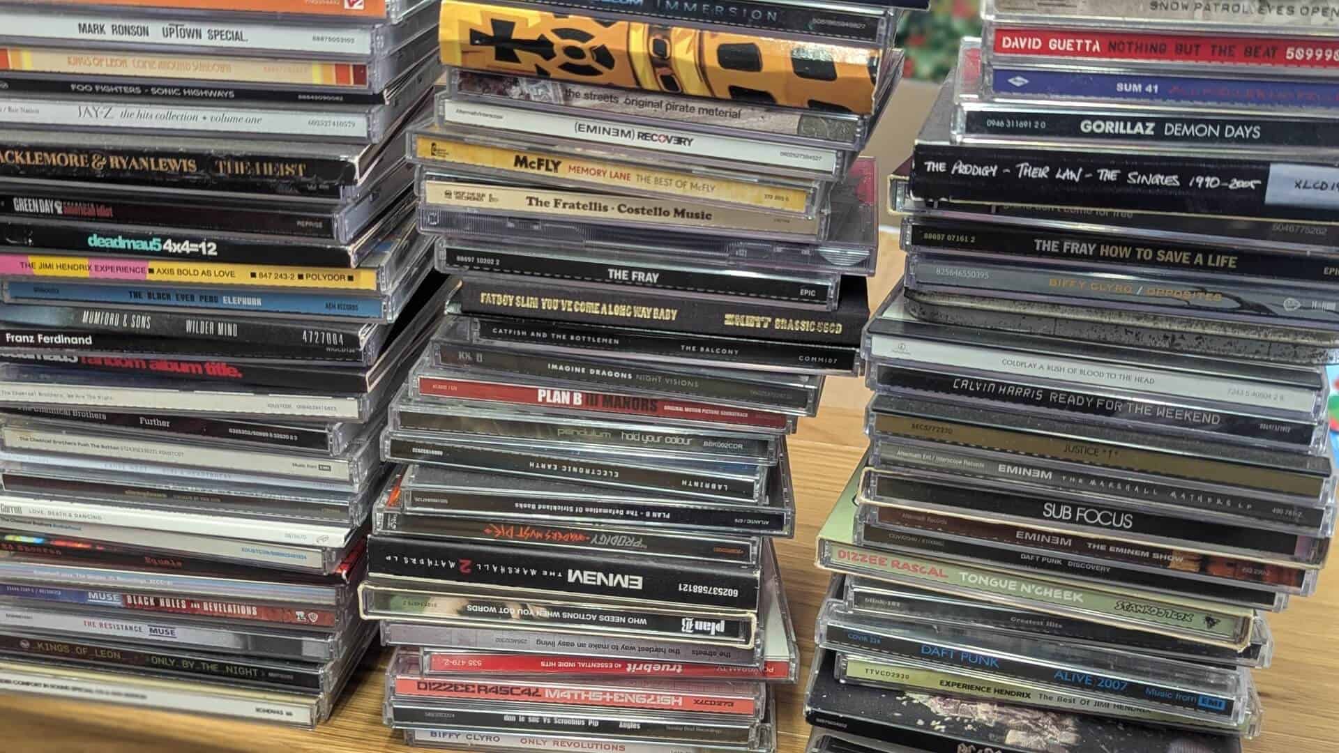 Multiple tall stacks of music CD jewel cases resting on a wooden table, showing spines of popular album titles.