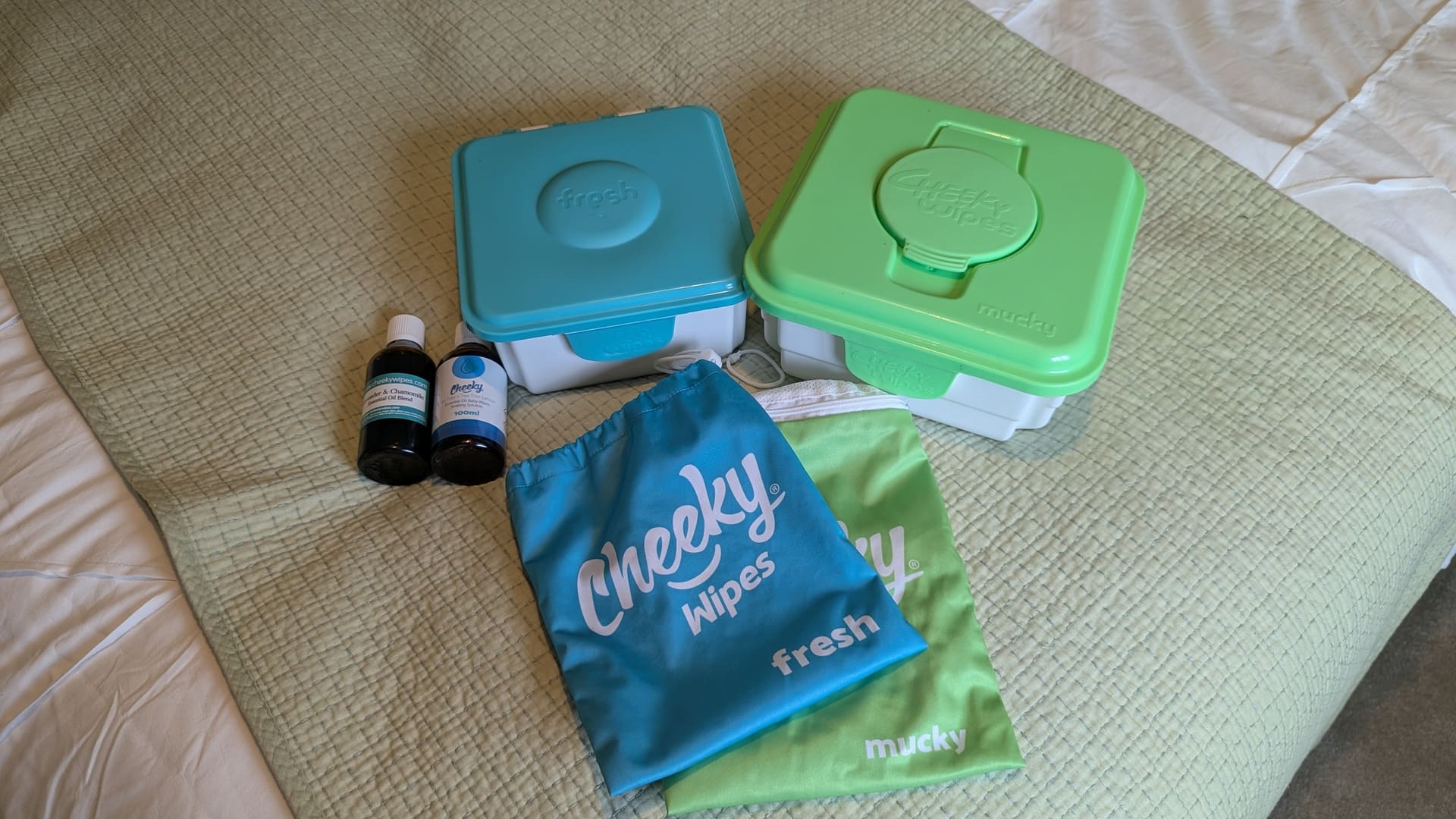 Cheeky Wipes Premium All-In-One Kit laid out on a bed featuring blue fresh tub, green mucky tub, essential oils, wet bags, and mesh laundry bag for an honest Cheeky Wipes review.