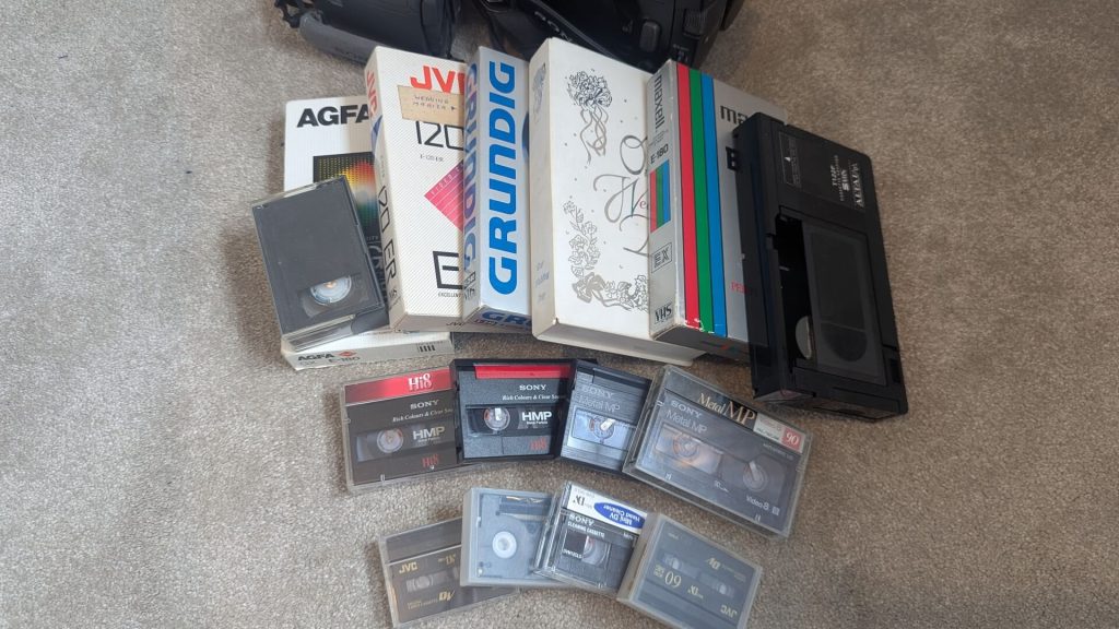 A collection of various video tape formats including full-size VHS, Hi8 cassettes, and MiniDV tapes scattered on a carpet.