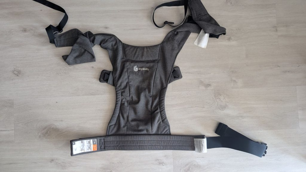 Ergobaby Embrace soft jersey baby carrier laid out flat on a light oak wooden floor showing straps and waist belt.