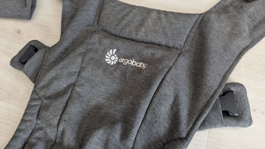 Close-up of the grey knit fabric and white embroidered logo on the front panel of an Ergobaby Embrace carrier.