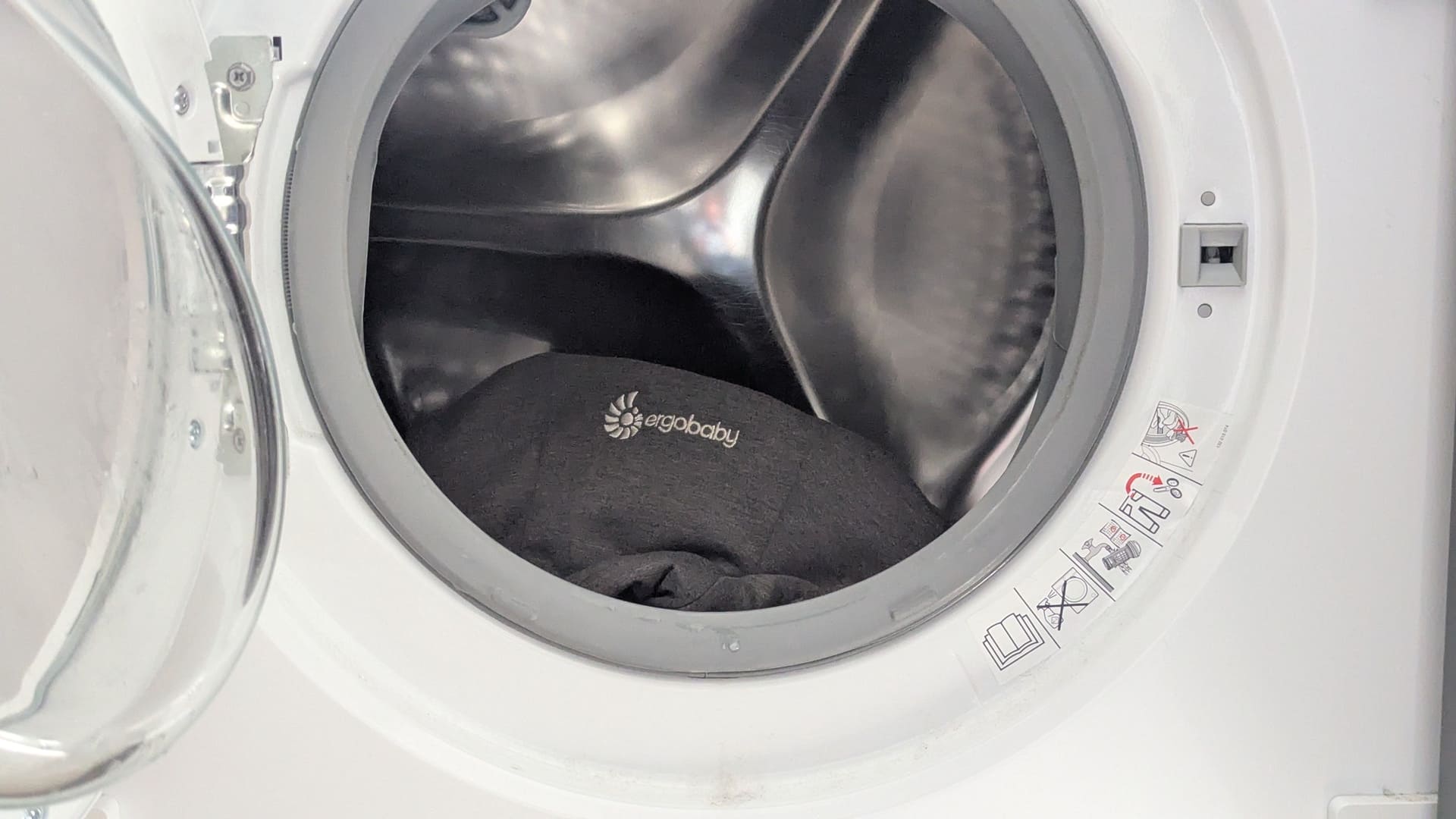 Charcoal grey Ergobaby Embrace baby carrier loaded into an open washing machine drum ready for a delicate wash.