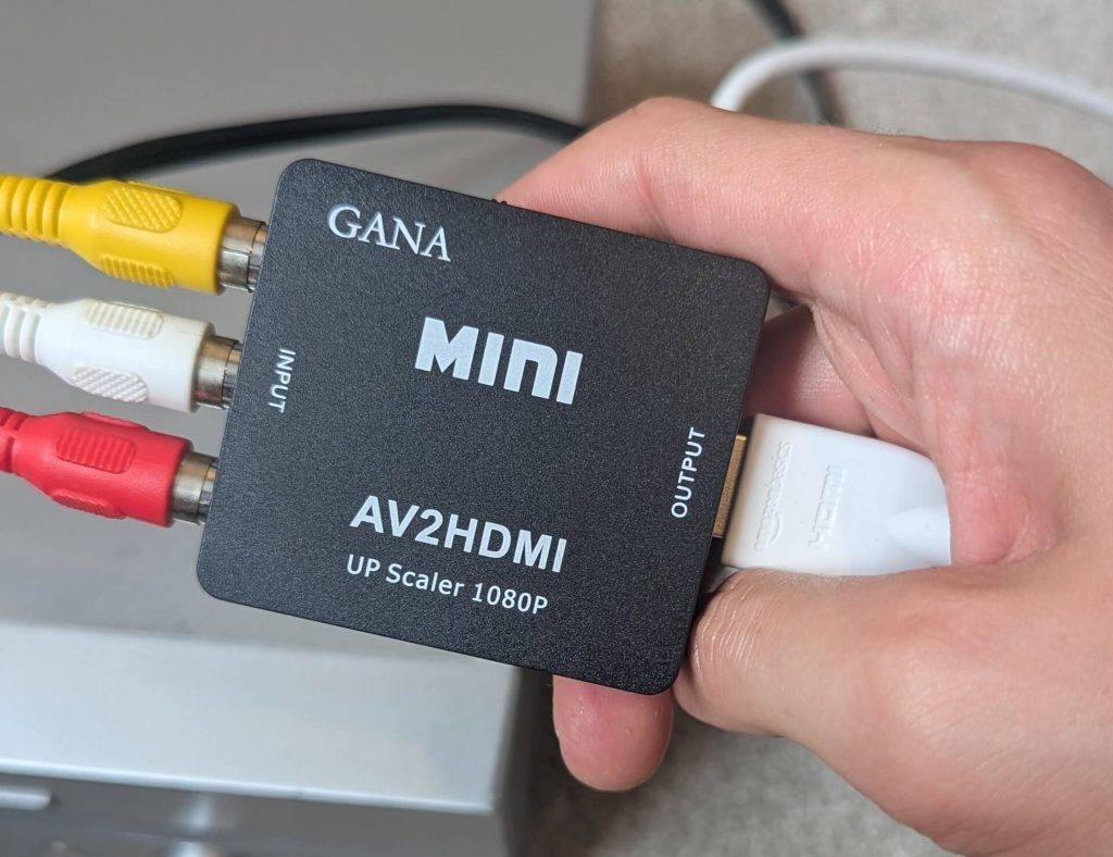 Hand holding a black GANA AV2HDMI Mini upscaler box with RCA inputs on the left and an HDMI output on the right.