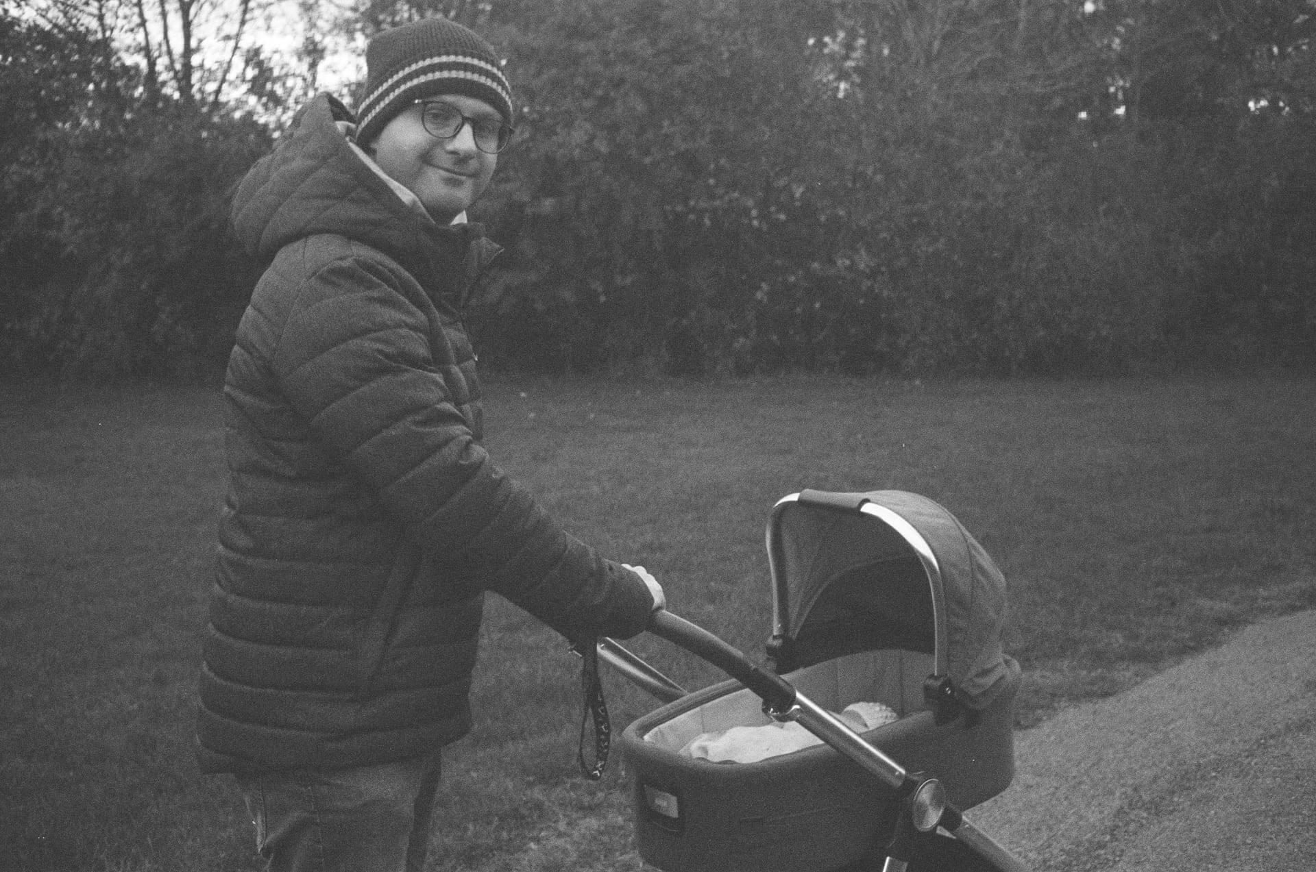 Dad, The North East Dad in a winter coat and bobble hat pushing a pram with a newborn baby outdoors on a park path.