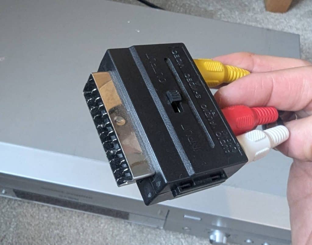 Hand holding a black SCART block adapter connected to yellow, red, and white RCA cables.