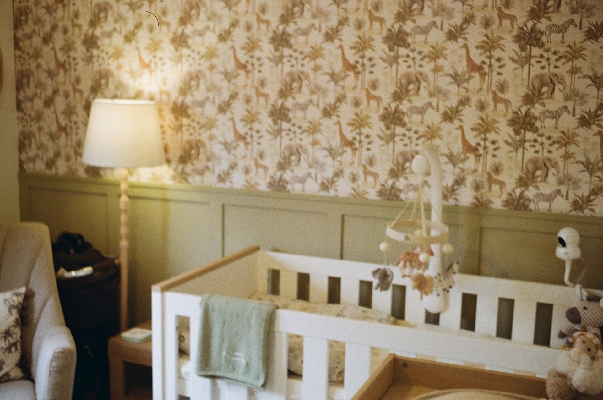 Cosy nursery corner featuring a white wooden cot with a mobile, standing lamp, and safari wallpaper during sleep training a 10 month old.
