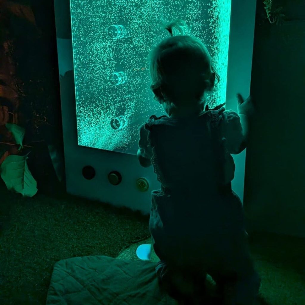 Silhouette of an 11-month-old baby girl standing up against a glowing green bubble wall in a dark baby sensory room.
