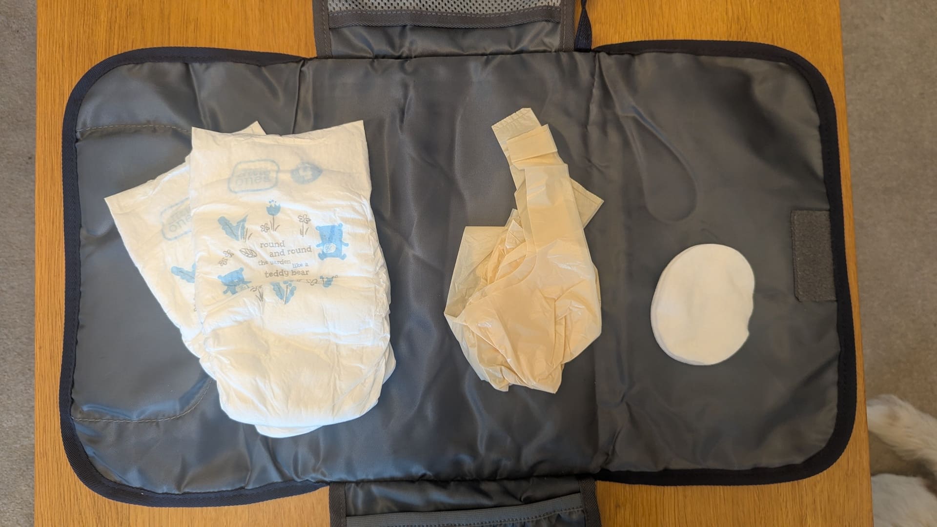 Open blue portable changing mat flat-lay showing three nappies, three nappy bags, and cotton wipes on a wooden table.