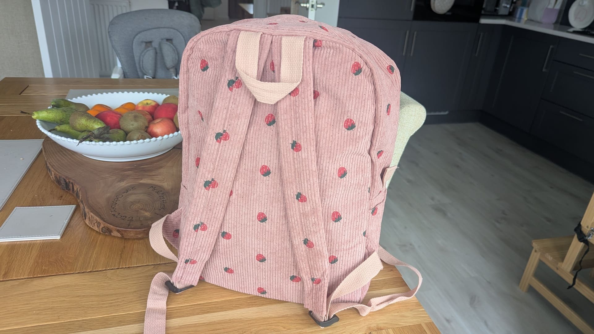 Pink corduroy nursery backpack with strawberry print sitting packed on a wooden dining table.