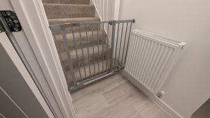 Hauck silver pressure-fit safety gate fitted across the bottom of a carpeted staircase next to a radiator.