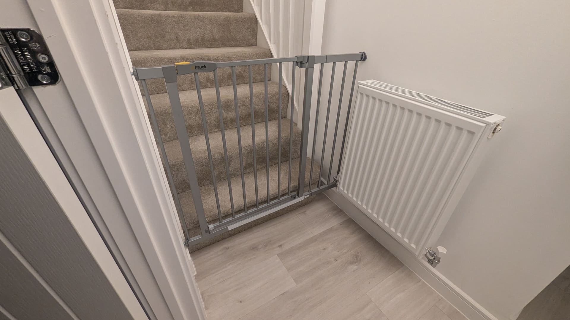 Hauck silver pressure-fit safety gate fitted across the bottom of a carpeted staircase next to a radiator.