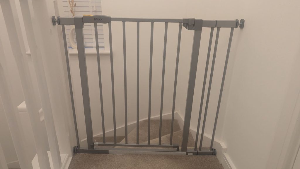 Silver Hauck pressure fit safety gate installed at the top of a carpeted staircase in a home.