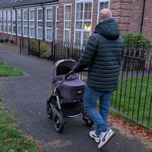 Dad pushing a Mamas and Papas Ocarro pram with carrycot attachment along a paved path past brick houses.