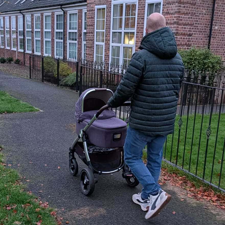 Dad pushing a Mamas and Papas Ocarro pram with carrycot attachment along a paved path past brick houses.