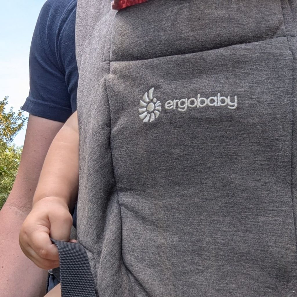 Close-up front view of grey Ergobaby Embrace baby carrier worn by dad outdoors on a trail.